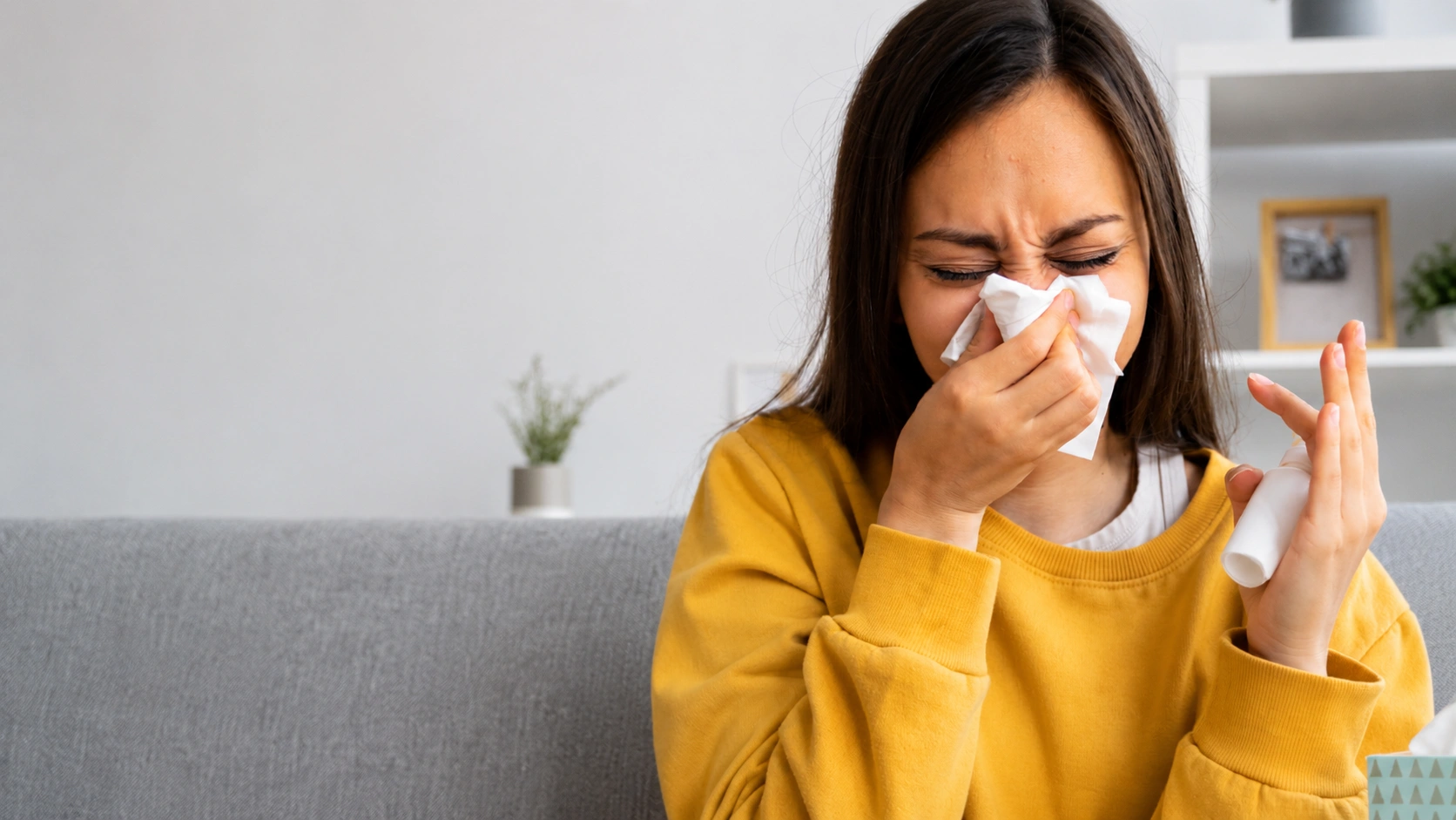 Donna che starnutisce coprendosi il naso con un fazzoletto di carta sul divano a casa. Visita allergologica a Varese.